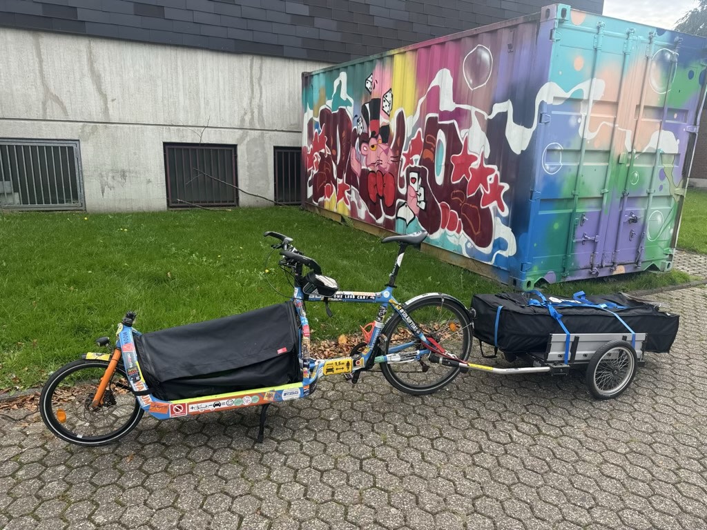Ein blaues Bullitt-Lastenrad mit geschlossener Transportbox und angehängtem Fahrradanhänger, beladen mit einer schwarzen Zeltasche, geparkt vor einer bunten Graffiti-Wand auf einem Steinpflasterplatz. A blue Bullitt cargo bike with a closed cargo box and an attached bicycle trailer loaded with a black tent bag, parked in front of a colorful graffiti wall on a paved courtyard.