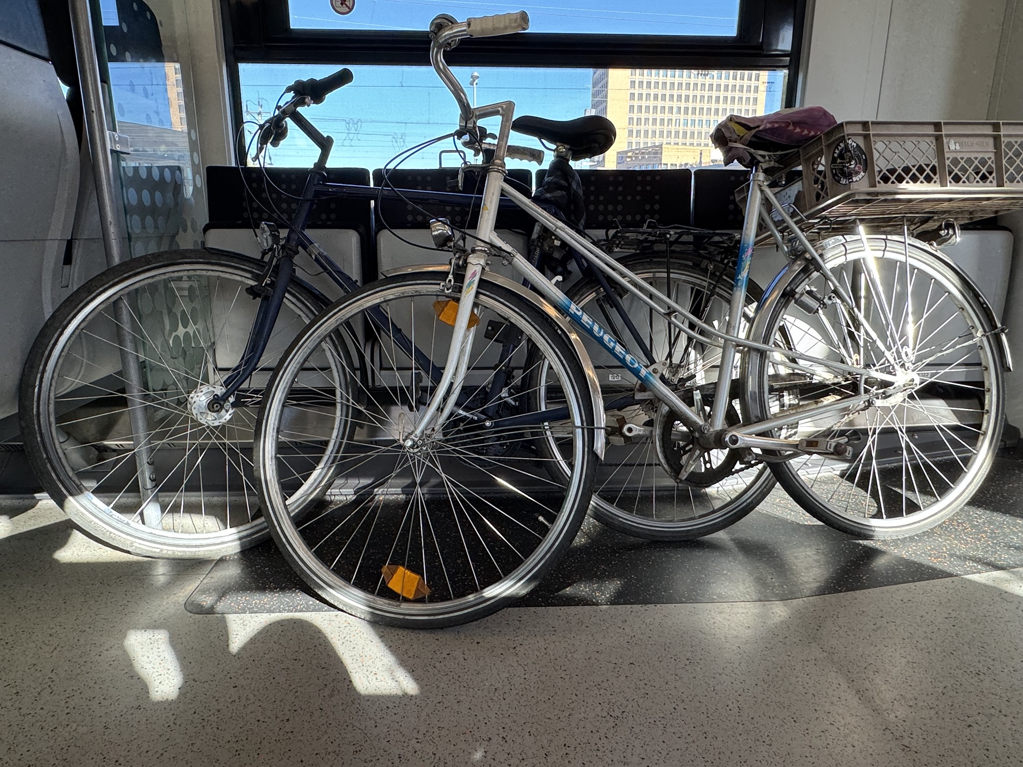 Ein altes silbernes Peugeot-Fahrrad mit GepÀcktrÀgerkiste steht neben einem blauen Fahrrad im Mehrzweckabteil des RE1.