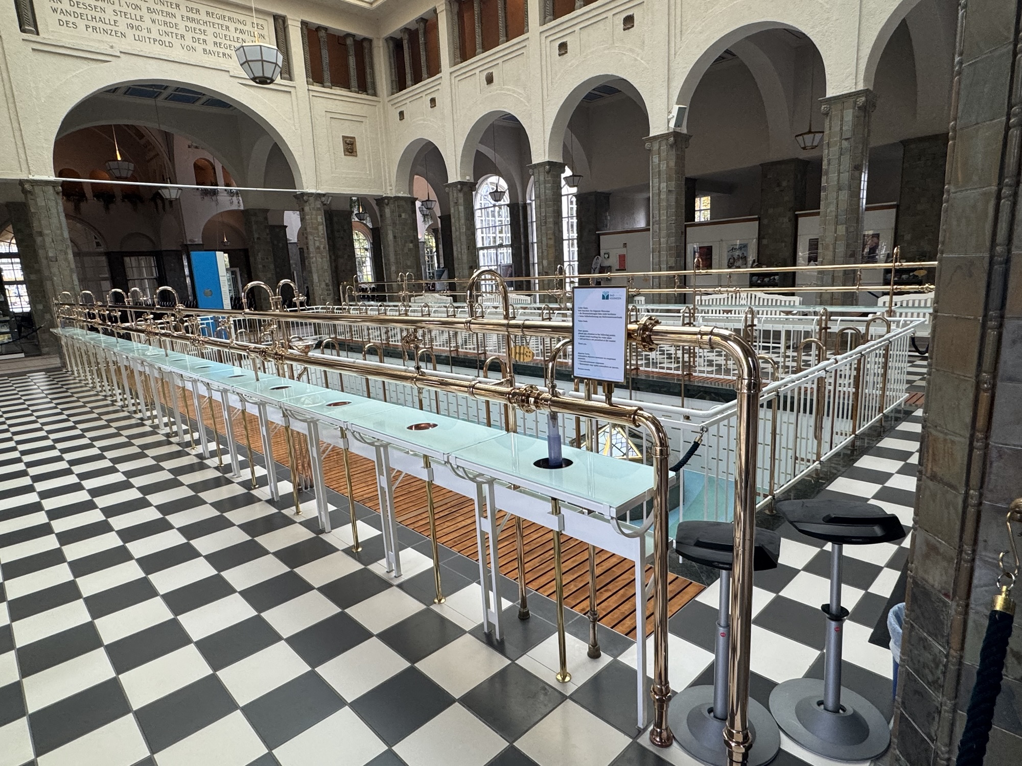 Historische Brunnenhalle der Wandelhalle Bad Kissingen mit langer Ausschank-Theke aus Messing und Glas vor schwarz-weiß gekacheltem Boden.