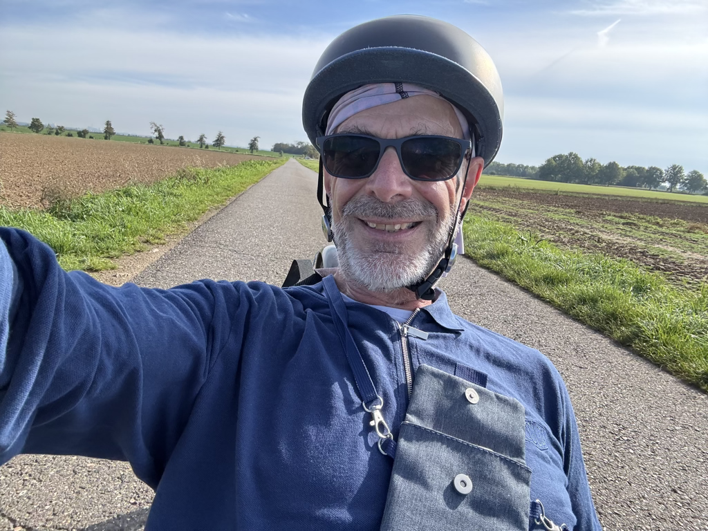 Selfie von Axel mit Fahrradhelm und Sonnenbrille entspannt im Liegedreirad auf einem Feldweg.