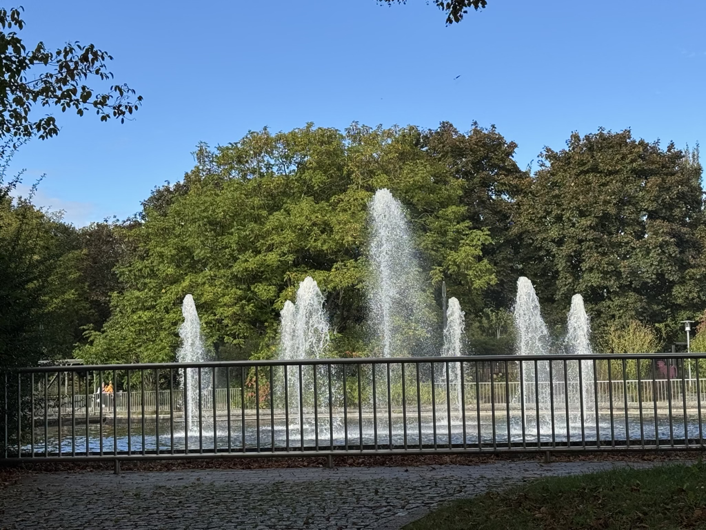 Hohe Fontänen einer Wasserspiel-Anlage hinter einem Geländer im Bürgerpark Langenfeld.